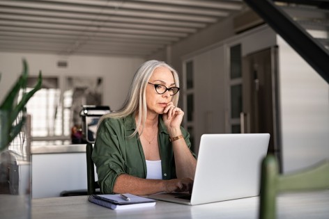 Eine etwa 60 jährige Frau sitzt an einem Tisch und arbeitet an einem Laptop. Neben ihr liegen ihre Notizen.