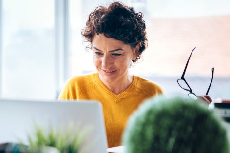 Eine brünette Frau hat eine Brille in der linken Hand, sitzt an einem Schreibtisch und schaut auf Ihren Laptop.