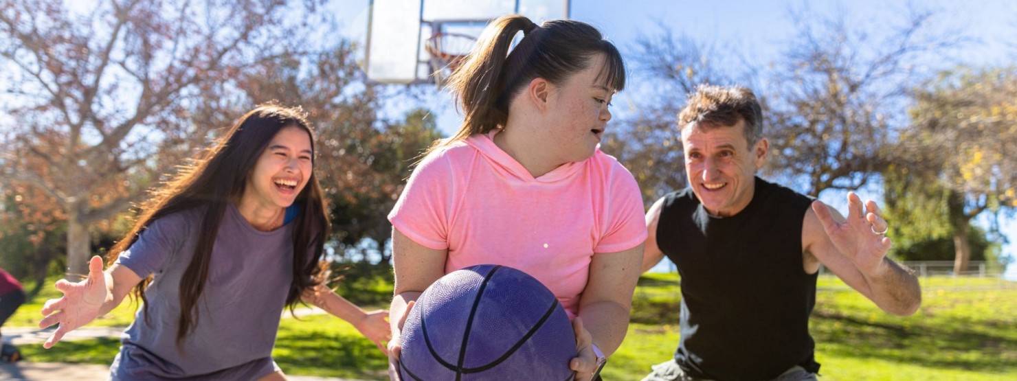Ein Mädchen mit Down-Syndrom spielt mit zwei weiteren Personen Basketball auf einem Platz im Freien.