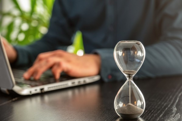 Eine Sanduhr steht auf dem Tisch. Im Hintergrund arbeitet ein Mann am Laptop.