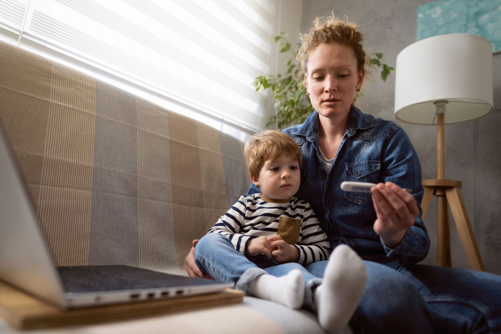 Eine Mutter sitzt mit ihrem kranken Kind auf der Couch und schaut besorgt auf ein Thermometer.