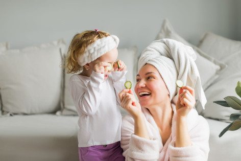 Eine etwa 30 jährige Frau befindet auf dem Boden vor einem Sofa. Sie trägt ein Handtuch als Turban und einen Bademantel. Gemeinsam mit ihrer etwa 3 jährigen Tochter halten sie sich Gurkenscheiben vor die Augen. Beide lachen ausgiebig.