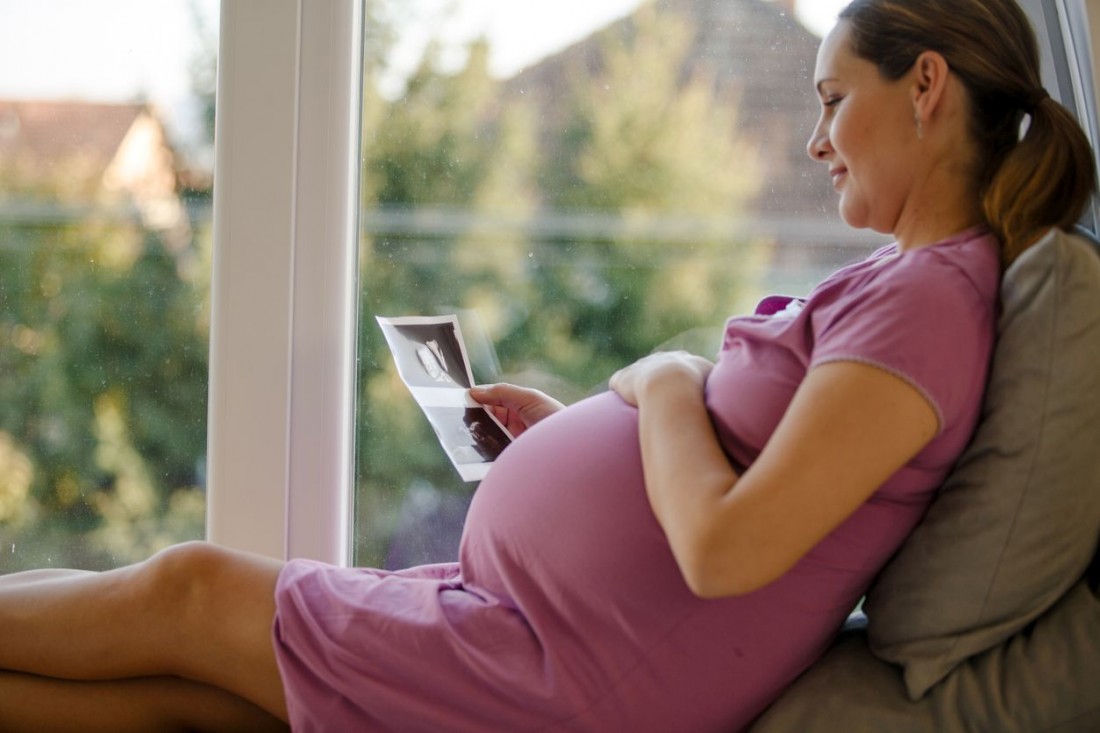Eine schwangere Frau sitzt am Fenster und betrachtet die Ultraschallbilder ihres Kindes.