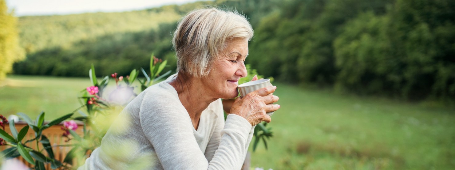 Eine Seniorin steht an einen Zaun gelehnt und trinkt eine Tasse Tee. Sie schaut in die Natur und lächelt.