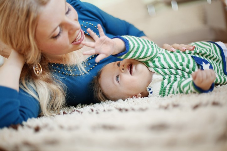 Eine etwa 30-jährige Mutter und ihr Säugling liegen auf einem Teppich und spielen miteinander.