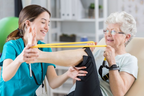Eine Seniorin führt eine Beinübung mit einem Gummiband aus. Dabei hilft ihr eine Physiotherapeutin.