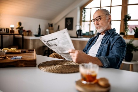 Ein etwa 60 jähriger Mann sitzt an einem Tisch und liest eine Zeitung. Er trägt eine Brille.