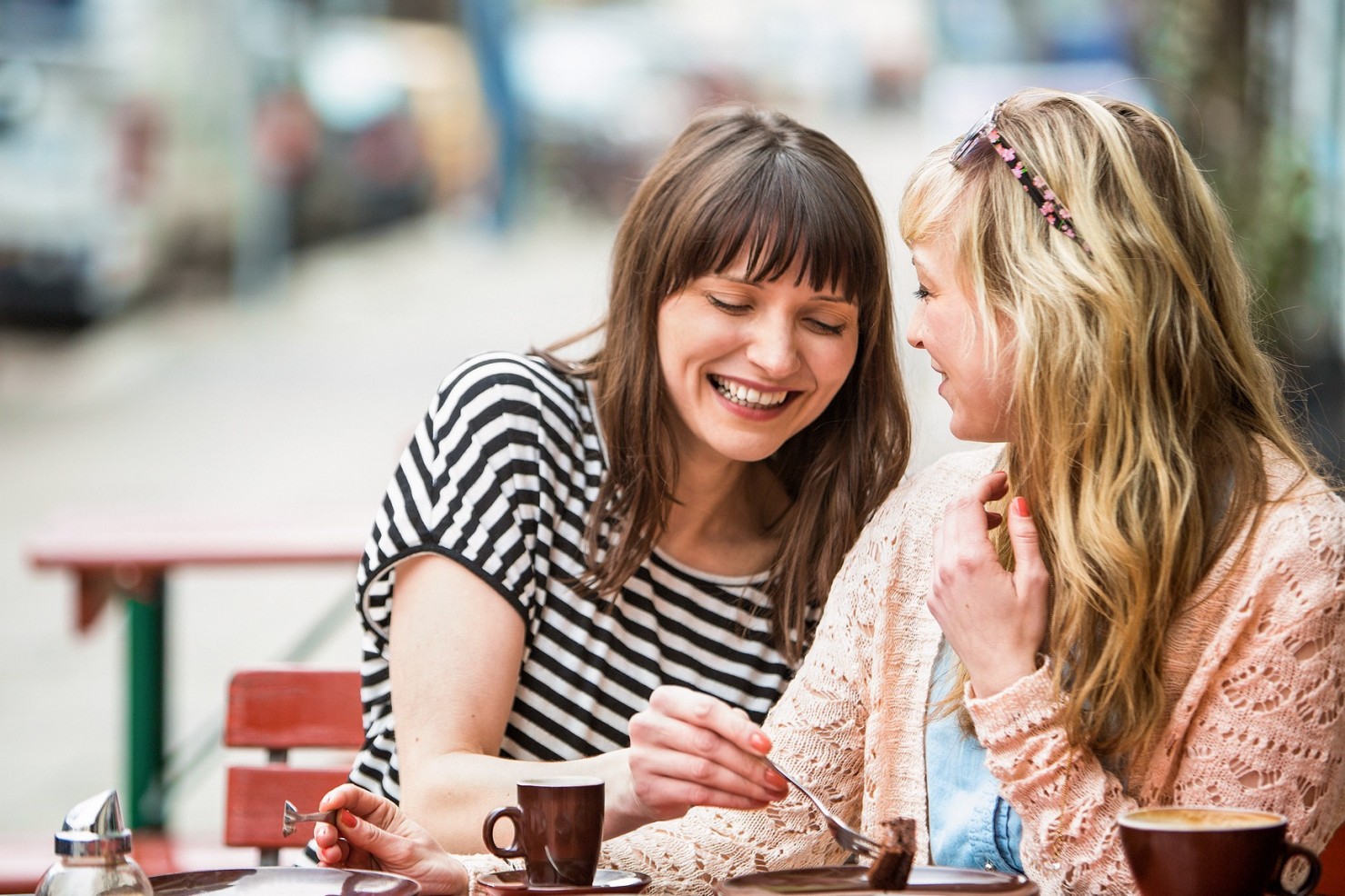 Zwei Freundinnen, beide circa 30 Jahre alt, sitzen draußen in einem Café. Eine der Frauen nimmt mit der Gabel das letzte Stück Brownie vom Teller ihrer Freundin.