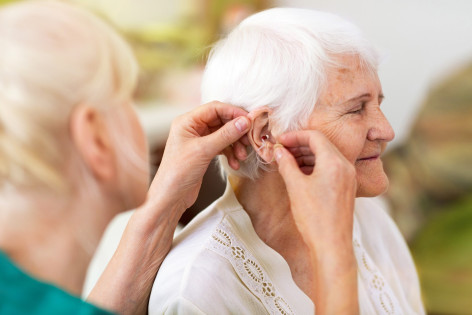 Einer Seniorin wird durch eine weibliche Person eine Hörhilfe in das Ohr eingesetzt.