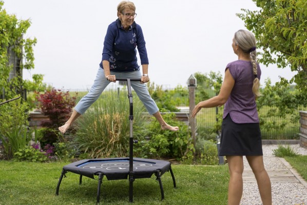 happy smiling fit senior woman retiree jumping in straddle on mini trampoline outdoors in garden, personel trainer female coach woman with enough social distance giving instructions in times of loosend coronavirus curfew