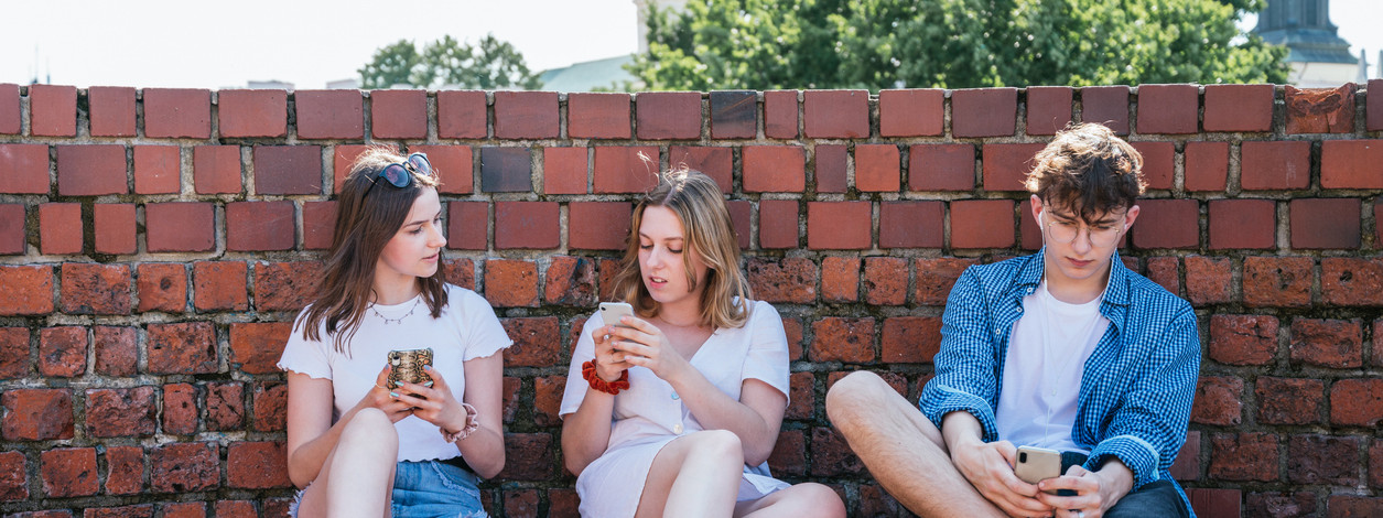Drei Jugendliche sitzen an einer Mauer und schauen süchtig auf ihre Smartphones.