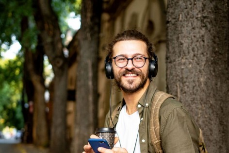 Eine etwa 40 jähriger Mann sitzt am Straßenrand einer Allee, hat Kopfhörer auf und sein Handy in der Hand. Dabei lächelt er.