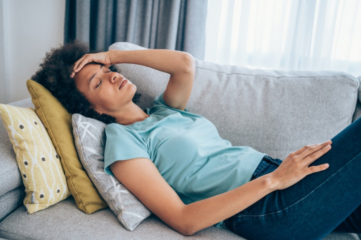 Junge Frau liegt auf dem Sofa und fasst sich an die Stirn wegen Kopfschmerzen.