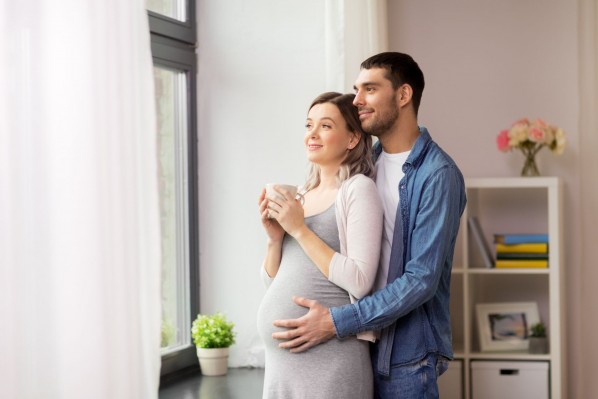 Ein Pärchen steht gemeinsam am Fenster und schaut heraus. Die Frau ist schwanger und hält eine Tasse in den Händen. Der Mann umarmt sie von hinten und berührt ihren Babybauch.