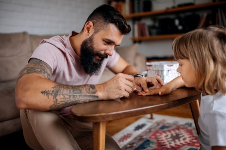 Ein etwa 30 jähriger Mann sitzt mit seiner Tochter gemeinsam am Wohnzimmertisch. Er bemalt ihre Fingernägel mit Nagellack für Kinder. Seinen Arm zieren mehrere Tattoos. 