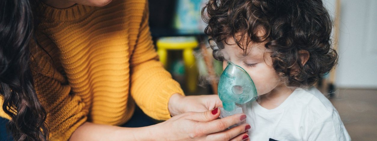 Eine Mutter hält ihrem etwa 2-jährigen Sohn ein Inhalator vor die Nase. Sie trägt einen gelben Pullover. 