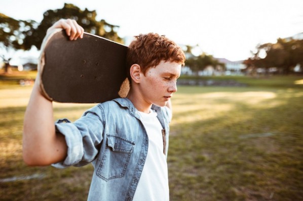 Ein etwa 12-jähriger Junge steht mit geschlossenen Augen auf einer Wiese. Er hält mit seinen Armen ein Skateboard hinter seinem Kopf.