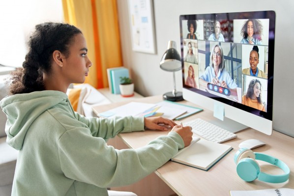 Eine etwa 20 jährige Frau sitzt im HomeOffice vor einem Computer und macht sich Notizen. Auf dem Computerbildschirm findet ein Meeting mit anderen Kollegen statt.