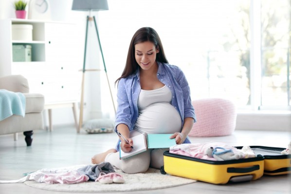 Eine etwa 30 jährige schwangere Frau sitzt in einem Wohnzimmer auf dem Boden. Sie schreibt etwas in ein Notizbuch. Um sier herum liegt ein geöffneter Koffer und Kleidung für die bevorstehende Geburt.