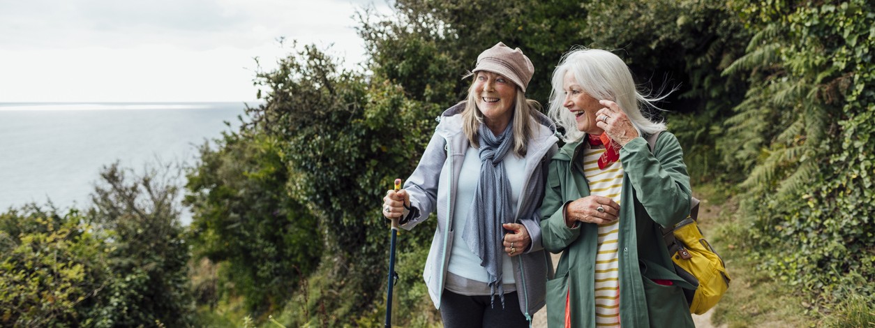 zwei etwa 60-jährige Frauen wandern an der Küste entlang. Im hIntergrund sieht man das Meer.