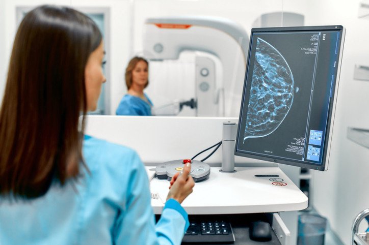 Eine Ärztin schaut auf ein Screeningbild einer Brust. Eine Patientin lässt im Hintergrund eine Mammographie durchführen.