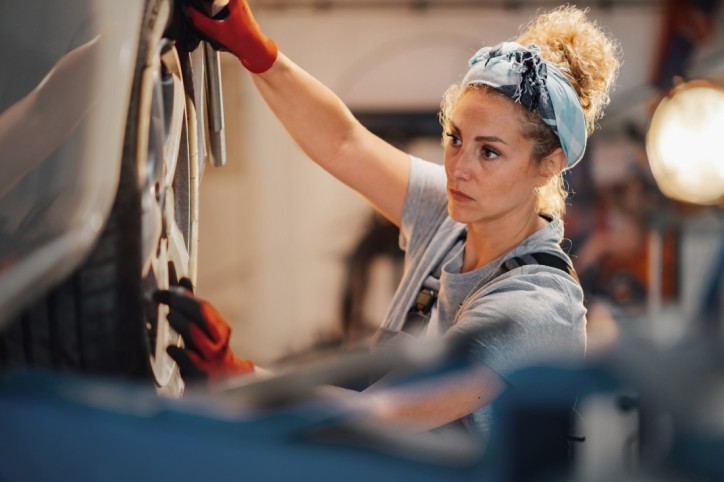 Eine etwa 40-jährige Automechanikerin wechselt an einem Auto ein Rad. Sie trägt Arbeitskleidung und rote Handschuhe. Sie befindet sich in einer Werkstatt bei künstlichem Licht.