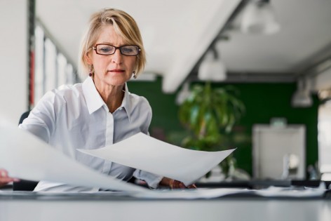 Eine etwa 50-jährige Frau mit blonden Haaren und Brille sortiert an einem Schreibtisch ihre Unterlagen.