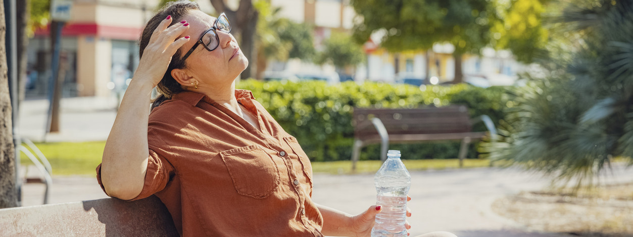 Eine Frau sitzt bei heißen Temperaturen auf einer Bank im Freien. Sie hält in der linken Hand eine Flasche Wasser und hat die Augen geschlossen.
