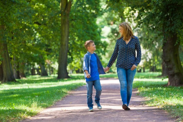 Eine Mutter läuft mit ihrem etwa 8 jährigen Sohn Hand in Hand durch einen grünen Park.
