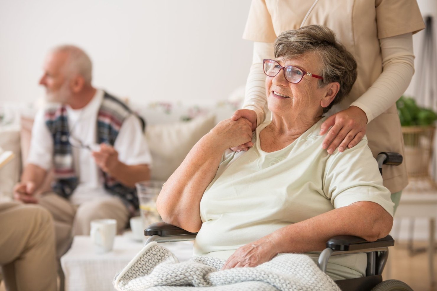 Eine Seniorin sitzt in einem Rollstuhl. Eine weibliche Person fasst ihr von hinten an die Schultern. Die Seniorin lächelt.