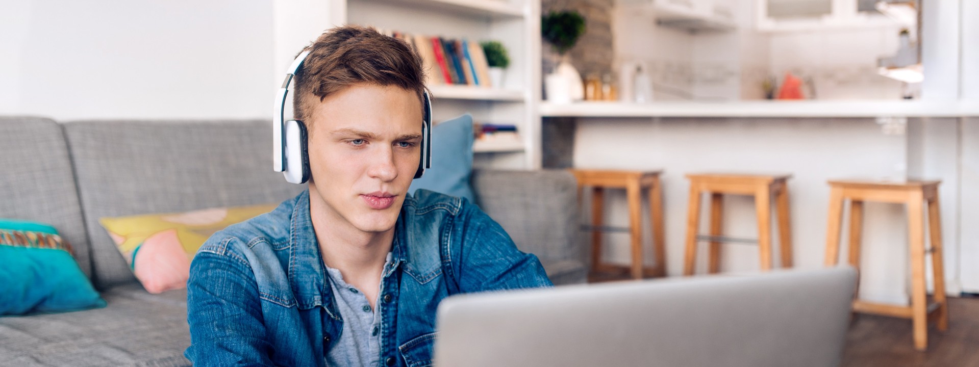 Ein circa 18-jähriger Mann mit Kopfhörern sitzt vor dem Sofa und liest auf seinem Laptop.