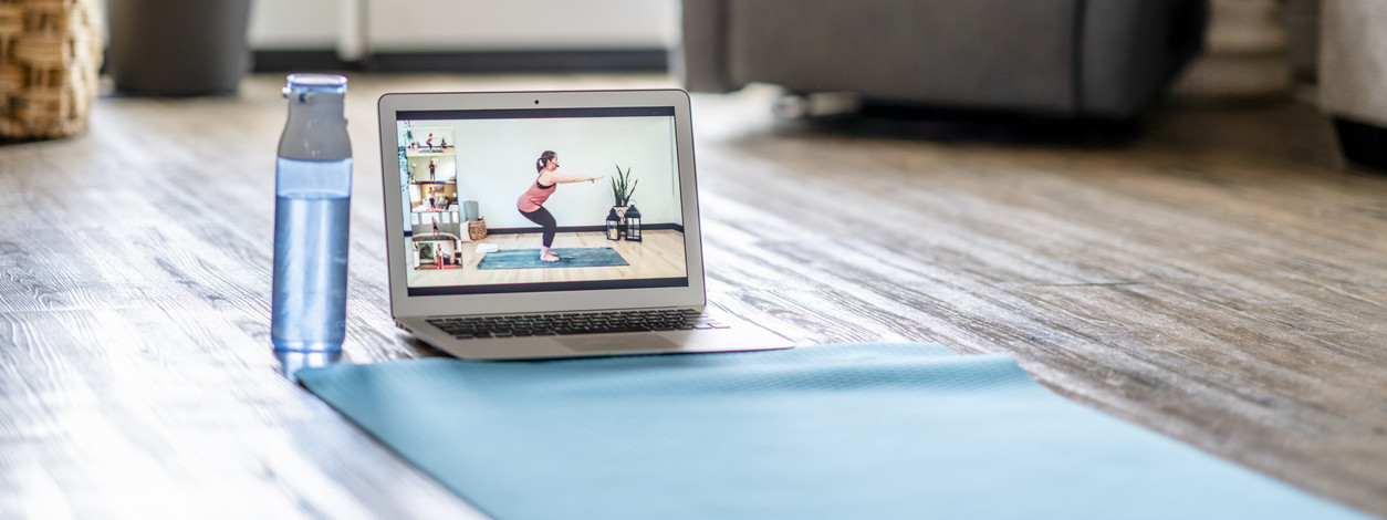 Auf dem Boden eines Wohnzimmers liegt eine Yogamatte und eine Wasserflasche. Auf dem Laptop-Bildschirm sieht man einen Online-Yoga-Kurs.