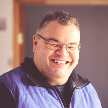 Nahaufnahme eines etwa 40-jährigen, leicht mehrgewichtigen Mannes mit kurzen Haaren und Brille. Er steht in einem Raum vor einer leeren Türöffnung. Er trägt eine violette Jacke und lächelt verschmitzt mit sichtbaren Zähnen und amüsiert zusammengekniffenen Augen.