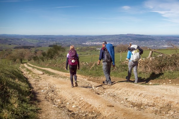 Drei Personen mit Rucksäcken laufen bei blauem Himmel auf einem Feldweg in Richtung Horizont.