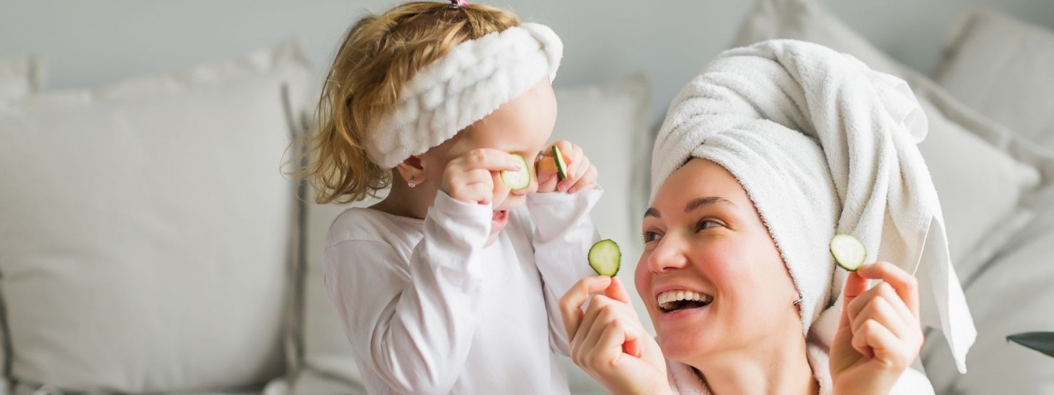 Eine etwa 30 jährige Frau befindet auf dem Boden vor einem Sofa. Sie trägt ein Handtuch als Turban und einen Bademantel. Gemeinsam mit ihrer etwa 3 jährigen Tochter halten sie sich Gurkenscheiben vor die Augen. Beide lachen ausgiebig.