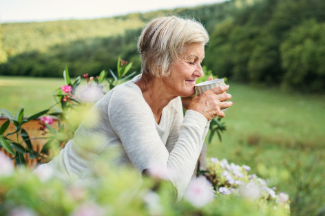 Eine Seniorin steht an einen Zaun gelehnt und trinkt eine Tasse Tee. Sie schaut in die Natur und lächelt.