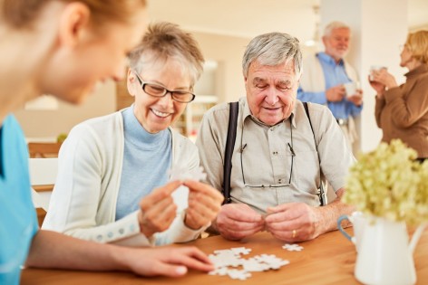 Eine Gruppe von Senioren sitzen um einen Tisch und puzzeln gemeinsam. Alle lachen ausgiebig.