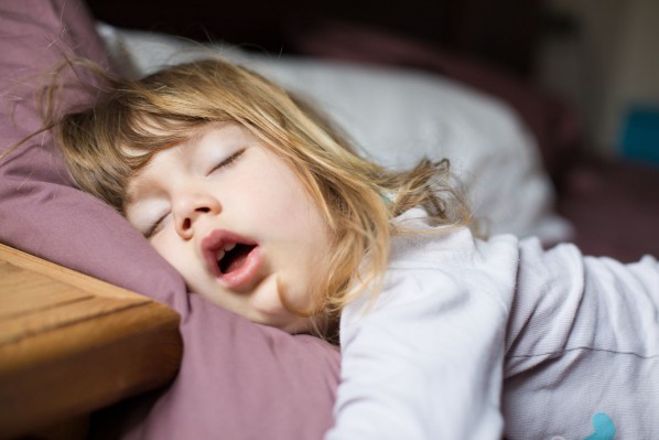 Ein etwa 6 jähriges Mädchen schläft in einem Bett. Den Mund hat sie dabei weit offen.