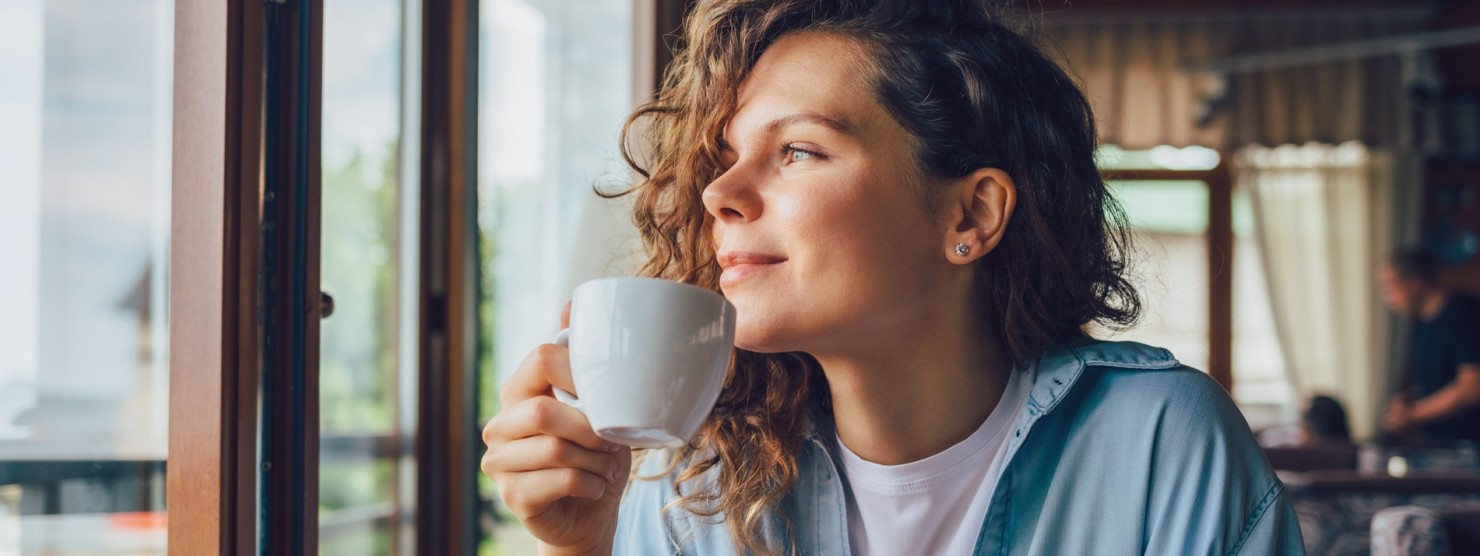 Etwa 25-jährige Frau schaut nachdenklich aus einem Fenster, während sie eine Tasse in der Hand hält.