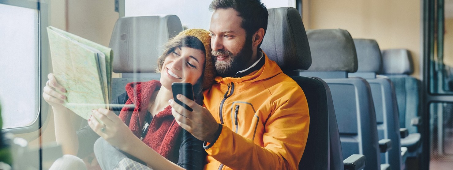 Reisendes Paar sitzt lächelnd im Zug und studiert die Route per Karte und Smartphone.
