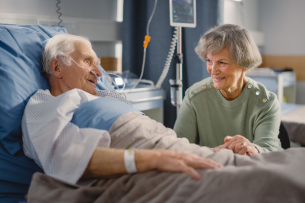 Ein älterer Patient liegt in einem Krankenhausbett und hat Besuch von einer Angehörigen. Er nimmt den Qualitätsvertrag zum postoperativen Delir in Anspruch.