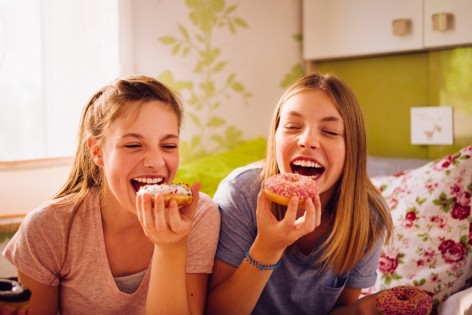 Zwei ca 12-jährige Mädchen sitzen im Kinderzimmer auf dem Bett und essen einen Donut