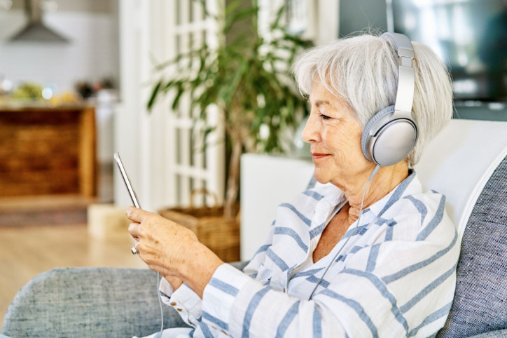 Eine Seniorin hört über ihre Kopfhörer den AOK Podcast Pflege. Dabei schaut sie auf ihr Smartphone und lächelt.