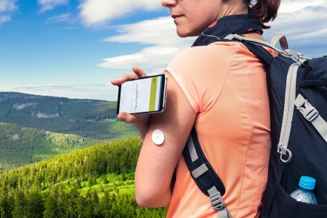 Frau mit Rucksack steht in den Bergen und misst mit einem Sensor und einem digitalen Lesegerät ihren Blutzucker