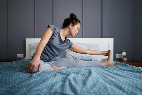 Eine etwa 25-jährige Frau macht auf ihrem Bett eine Dehnungsübung für die Beine.