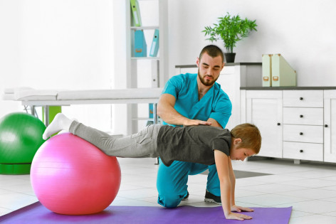Ein Junge turnt auf einem rosa Gymnastikball und wird dabei von einem Therapeuten unterstützt.