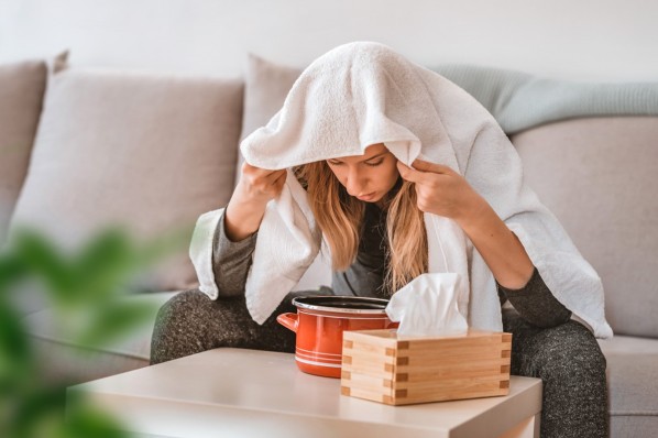 Junge Frau sitzt zu Hause auf ihrem Sofa und beugt sich zum Inhalieren über einen roten Topf. Auf dem Kopf hat sie ein weißes Handtuch hängen. Sie hält es mit den Händen fest.