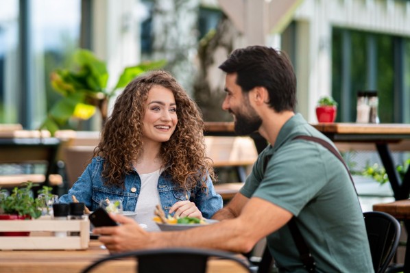 Eine etwa 25 jährige Frau und Mann sitzen in einem Cafe im Außenbereich und reden miteinander. Beide lächeln.
