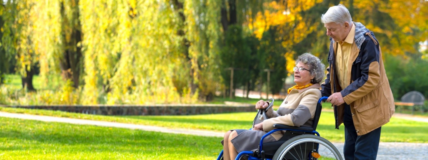 Ein Seniorenpaar befindet sich in einem grünen Park. Die Frau sitzt im Rollstuhl, der Mann steht hinter dem Rollstuhl und schiebt sie.