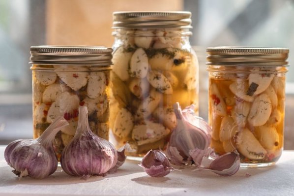 Auf einem Esstisch stehen drei Einmachgläser mit eingelegtem Knoblauch. Daneben liegen einzelne Knoblauchzehen.